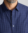 Man wearing a blue and navy plaid button-down shirt, showing the collar, chest area, and part of his beard against a neutral gray background.