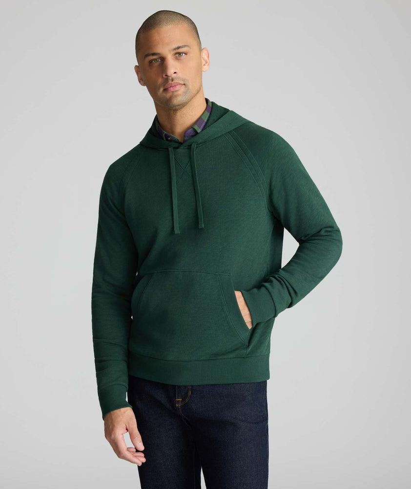 A man wearing a dark green pullover hoodie with drawstrings and a front kangaroo pocket, paired with dark jeans, against a solid light gray background.