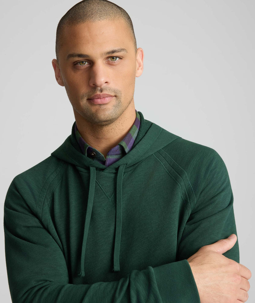 A man wearing a dark forest green hooded sweatshirt over a purple and green plaid button-down shirt, photographed against a solid light gray background.