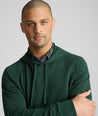 A man wearing a dark forest green hooded sweatshirt over a purple and green plaid button-down shirt, photographed against a solid light gray background.