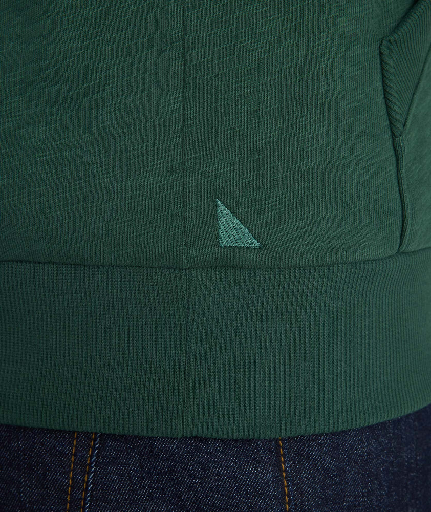 Close-up of a dark green ribbed knit garment with a small embroidered light green triangular logo, shown against dark blue jeans.