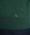 Close-up of a dark green ribbed knit garment with a small embroidered light green triangular logo, shown against dark blue jeans.