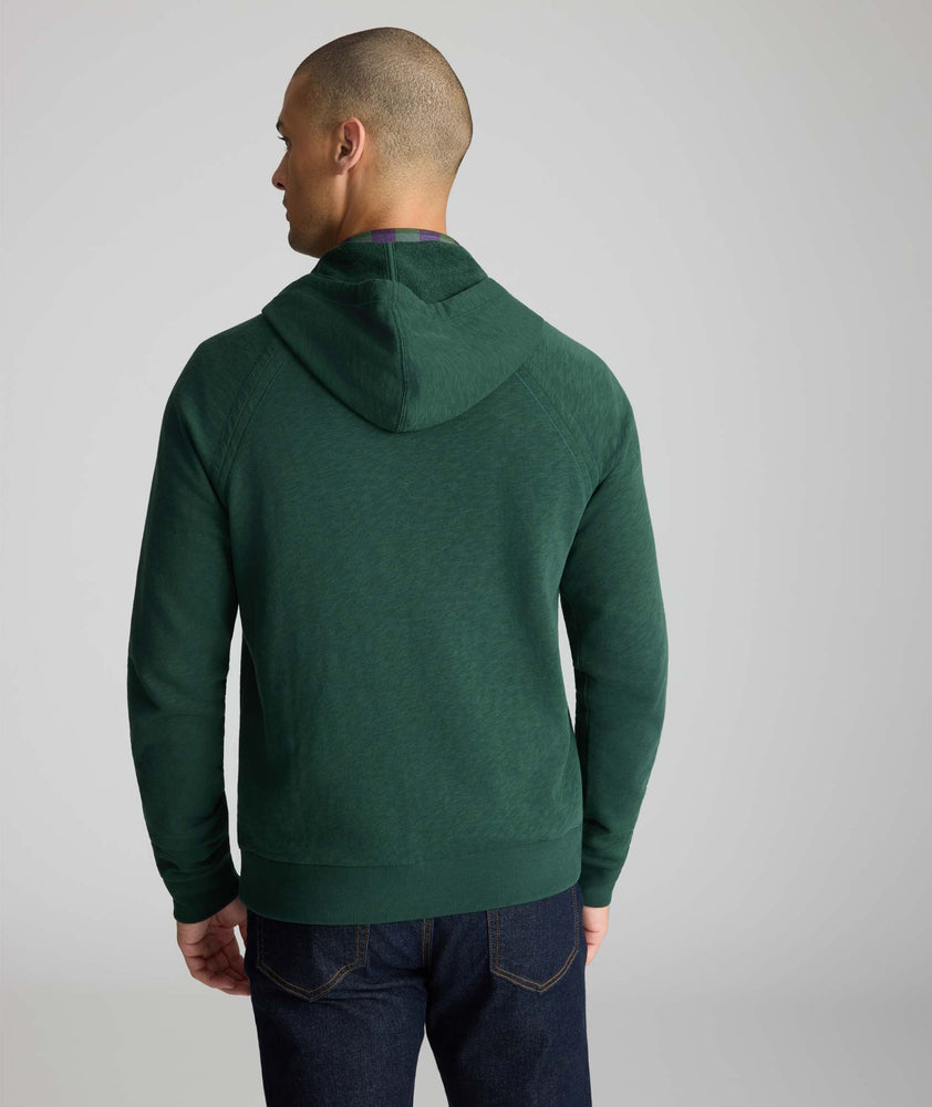 A man viewed from the back wearing a dark green pullover hoodie with raglan sleeves and dark blue jeans, set against a plain light gray studio background.
