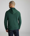 A man viewed from the back wearing a dark green pullover hoodie with raglan sleeves and dark blue jeans, set against a plain light gray studio background.