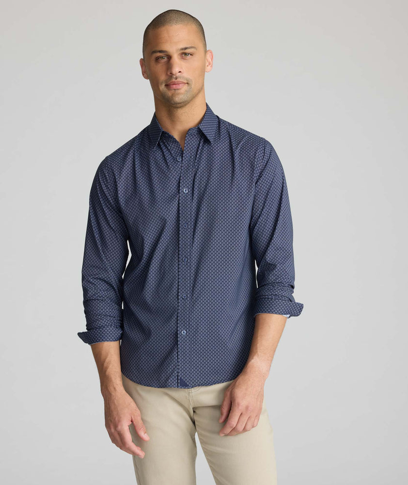 A man wearing a navy blue long-sleeve button-down shirt with a small white geometric pattern, paired with beige trousers, photographed against a neutral light gray background.