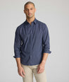 A man wearing a navy blue long-sleeve button-down shirt with a small white geometric pattern, paired with beige trousers, photographed against a neutral light gray background.