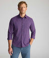 A man wearing a purple long-sleeve button-down shirt with a subtle grid pattern, paired with slate blue trousers, photographed against a neutral gray studio background.