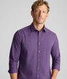 A man wearing a purple long-sleeve button-down shirt with a fine white grid pattern, photographed against a neutral light gray background.
