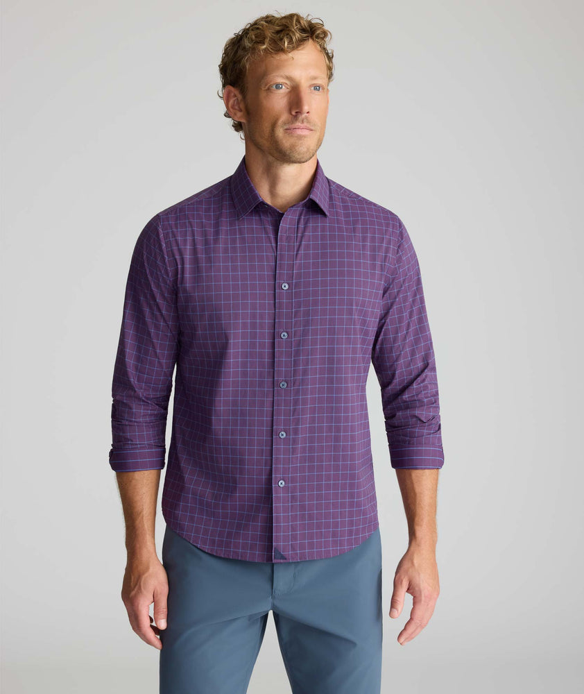 A man wearing a purple long-sleeve button-down shirt with a subtle blue grid pattern, paired with slate blue pants, standing against a light gray studio background.