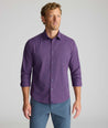 A man wearing a purple long-sleeve button-down shirt with a subtle blue grid pattern, paired with slate blue pants, standing against a light gray studio background.