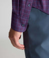 Close-up of a person wearing a purple and blue plaid button-down shirt with dark gray trousers, showing the cuff and hand.