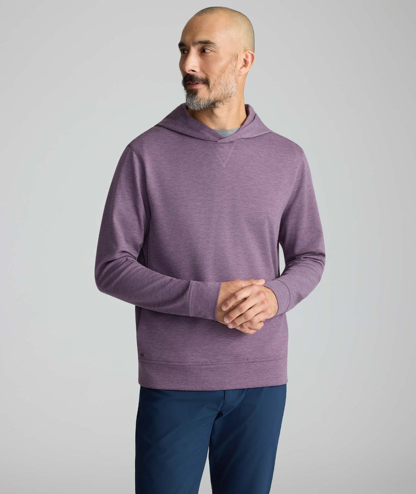 A man wearing a heathered purple long-sleeve hoodie and navy blue pants, standing against a light gray studio background.