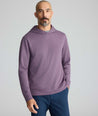 A man wearing a heathered purple long-sleeve pullover hoodie with a crew neck and hood, paired with navy blue pants, photographed against a solid light gray studio background.